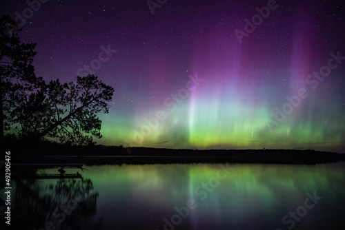 Northern lights erupt above a lake in Northern Minnesota in the dark sky overhead shining a rainbow of colors and brilliant light over the forests and water