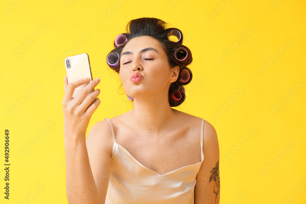 Beautiful young woman in hair curlers taking selfie on yellow background Beautiful young woman in hair curlers taking selfie on yellow background