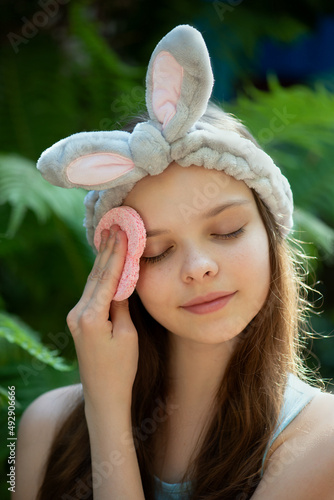 Portrait of cute little teenage girl enjoying skin care procedures. Beautiful little kid with funny bunny headband cleansing her face by heart shape sponge