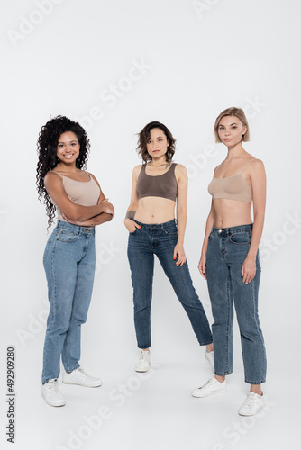 Young interracial women in tops and jeans posing on grey background.