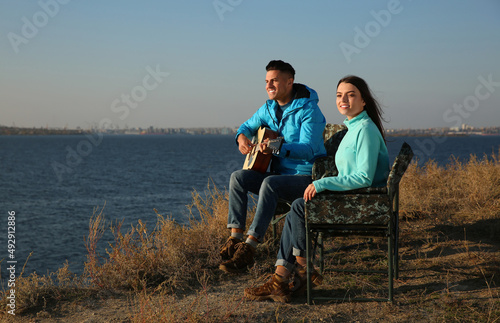 Couple with guitar resting in camping chairs near river on sunny day © New Africa