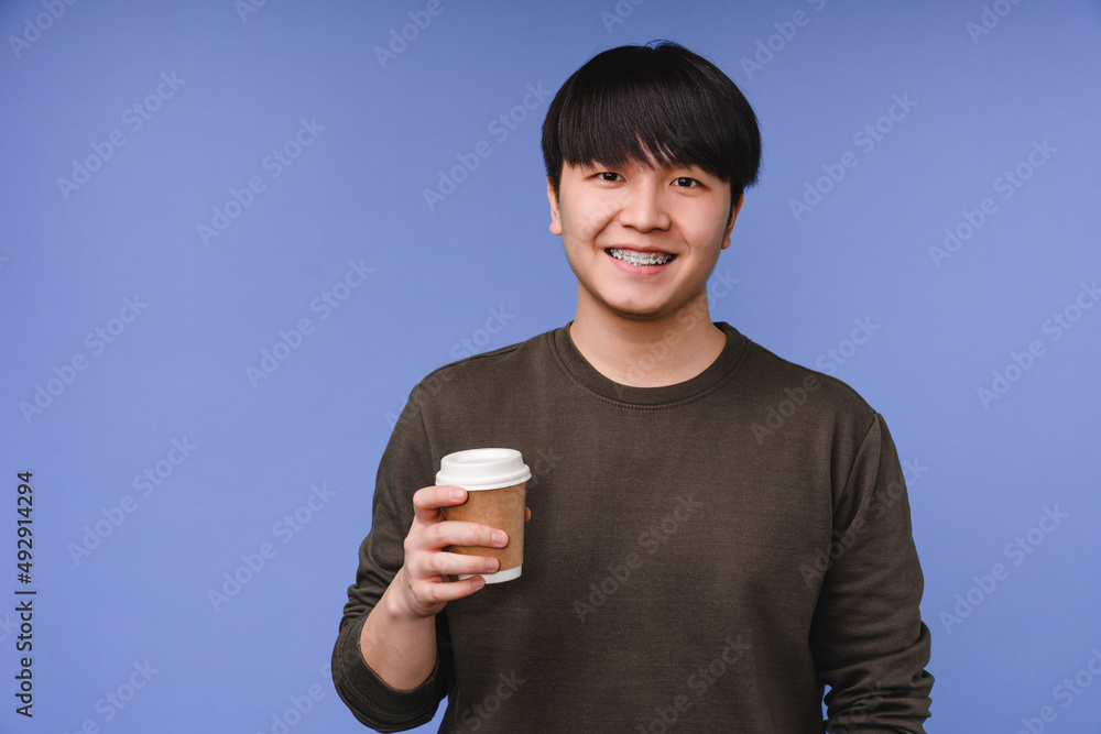 Young asian korean teenage boy man student drinking hot beverage, decaf coffee tea in paper cup isolated on blue background Young asian korean teenage boy man student drinking hot beverage, decaf coffee tea in paper cup isolated on blue background