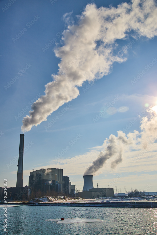 Manufacturing facility on water with two large silos releasing smog into air Manufacturing facility on water with two large silos releasing smog into air