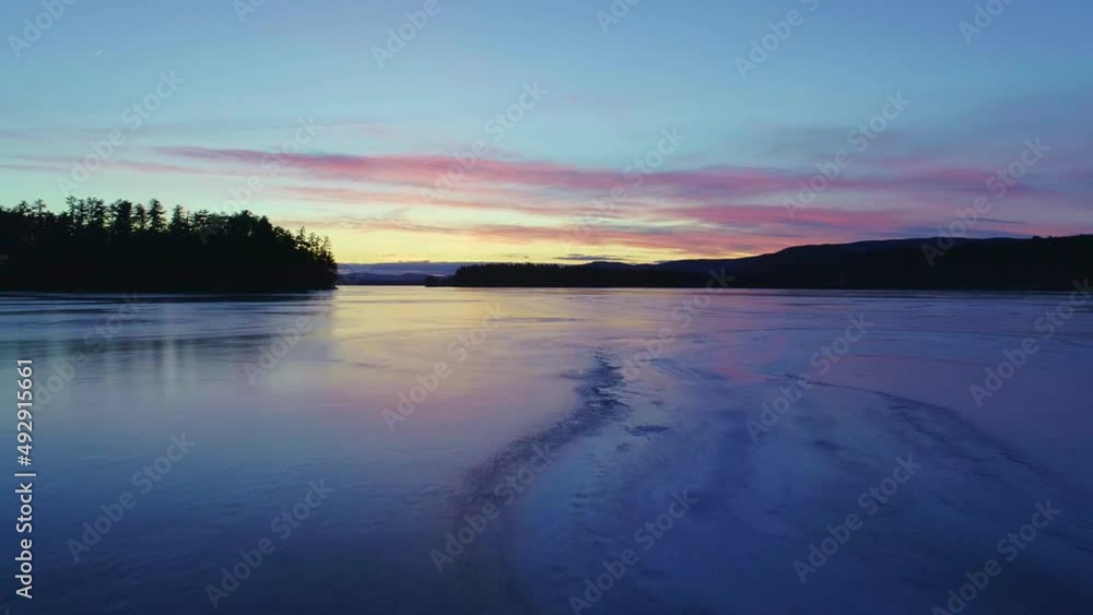 Drone shot flying low and slow over a frozen lake without snow toward the western sky just after sunset. It follows the crack in the ice and shows bubbles and other ice formations. Drone shot flying low and slow over a frozen lake without snow toward the western sky just after sunset. It follows the crack in the ice and shows bubbles and other ice formations.
