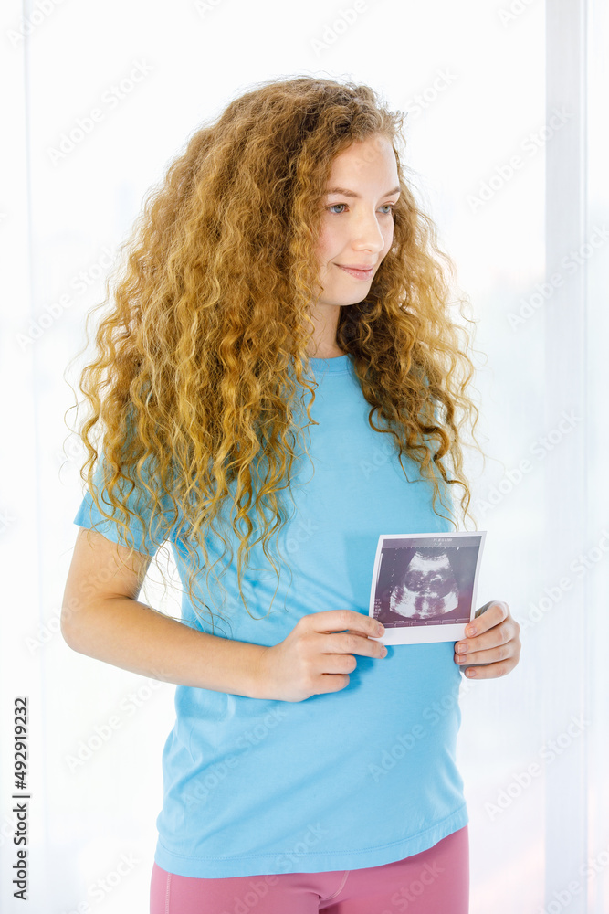 Caucasian young beautiful long curly hairstyle healthy pregnant mother in casual outfit standing holding showing baby ultrasound Xray film picture sonogram in hands in front of curtain background Caucasian young beautiful long curly hairstyle healthy pregnant mother in casual outfit standing holding showing baby ultrasound Xray film picture sonogram in hands in front of curtain background