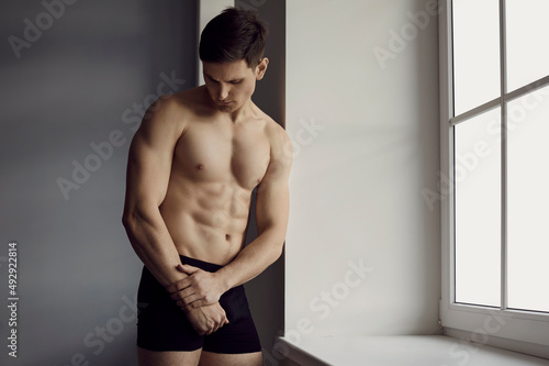 Portrait of young man with perfect athletic body with six-pack huge muscles in black classic underpants. Male model with wearing comfortable cotton underpants standing in bright daylight near window.