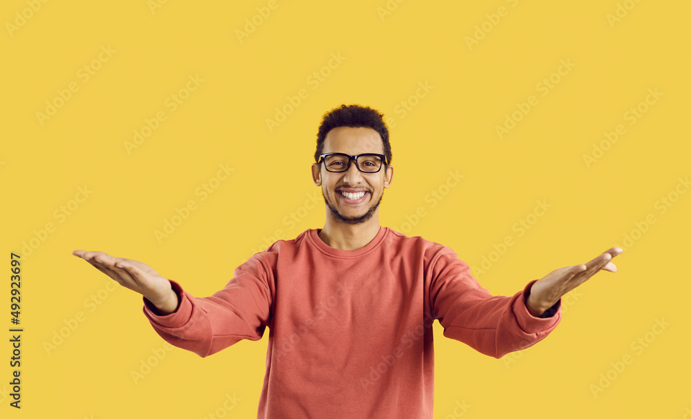 You did it. Congratulations. Happy cheerful positive young ethnic man in eyeglasses standing against yellow color background looking at camera, smiling and spreading his arms wide open for a warm hug - obrazy, fototapety, plakaty 