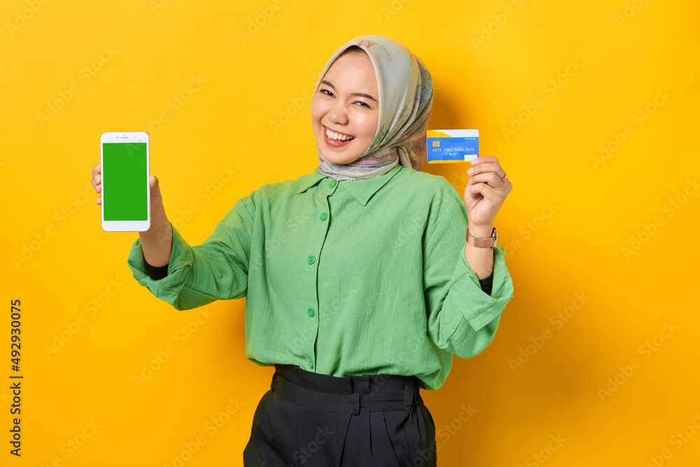 Cheerful young Asian woman in a green shirt showing blank screen smartphone and holding credit card on yellow background Cheerful young Asian woman in a green shirt showing blank screen smartphone and holding credit card on yellow background