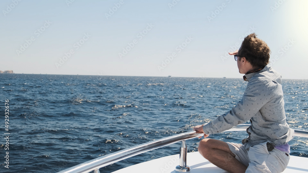 Man on yacht looks in distance, sunny morning. White sailing boat on blue sea waves, handsome man in light clothes, sunglasses sitting on bow. Luxury voyage summer travel adventure. Nautical lifestyle