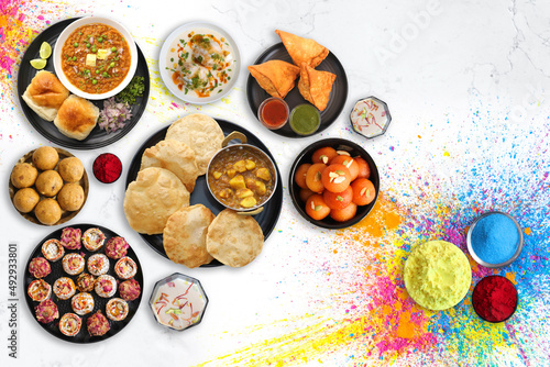 Assorted Indian Holi festival food like samosa, kachori, Puri bhaji, Pav Bhaji, Laddo, Sweets, Dahi vada and Almond milk Thandai drink with colorful background and colors with copy space.