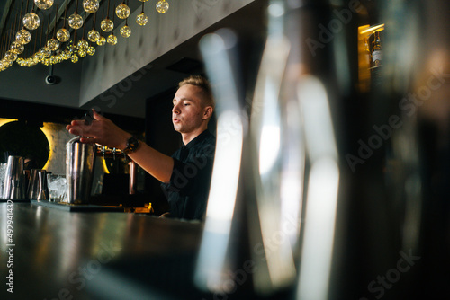 Side low-angle view of confident barman making refreshing alcoholic cocktail standing behind bar counter in modern dark nightclub, on background of shelves with different alcoholic drinks