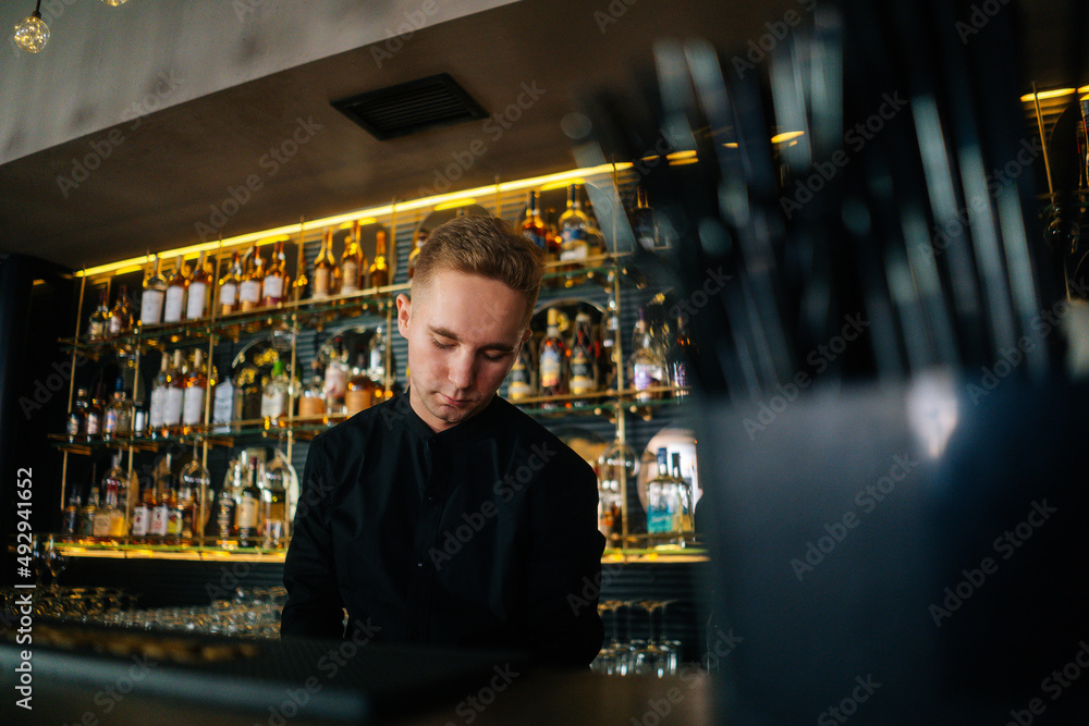 Low-angle view of confident bartender male making cocktail standing behind bar counter in modern dark nightclub, on blurred background of shelves with different alcoholic drinks, selective focus. Low-angle view of confident bartender male making cocktail standing behind bar counter in modern dark nightclub, on blurred background of shelves with different alcoholic drinks, selective focus.