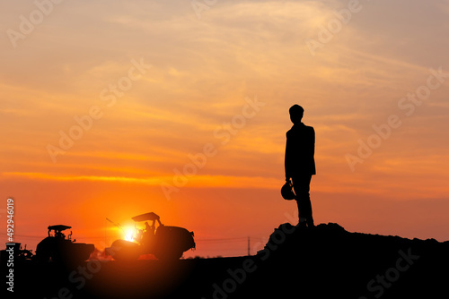 Silhouette of Engineer man with clipping path checking project, Foreman worker looking construction machinery in the road construction site sunset background