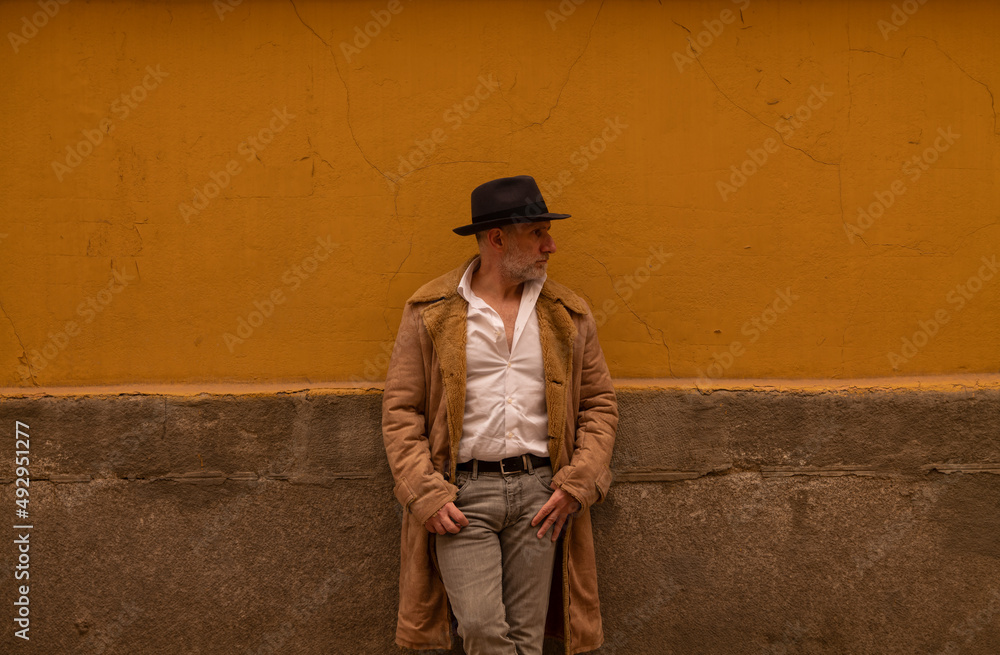 Portrait of adult man in hat and coat against yellow wall on street