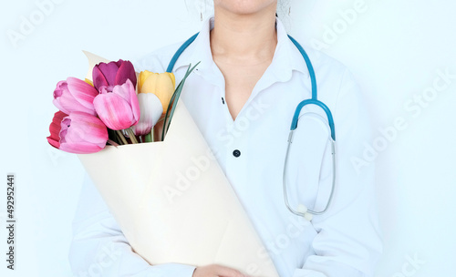 Doctor woman of Asian appearance, in white medical clothes with a stethoscope around her neck, holds a bouquet of flowers red tulips in her hands. The concept of congratulations on the day of the medi