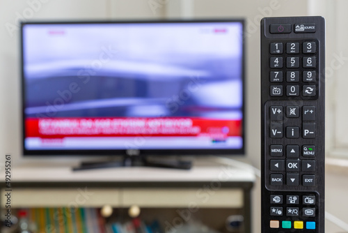 TV remote control in selective focus. The TV in the blurred background has a breaking news TV channel.