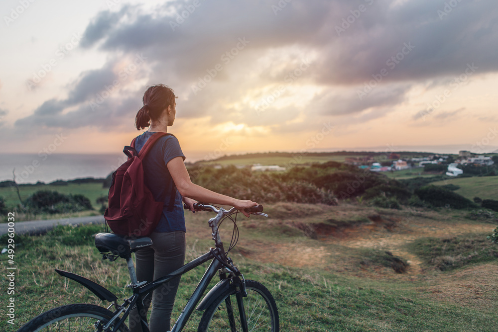 Fototapeta premium Young woman in the evening and watching the sunset while standing with her bicycle on a hill