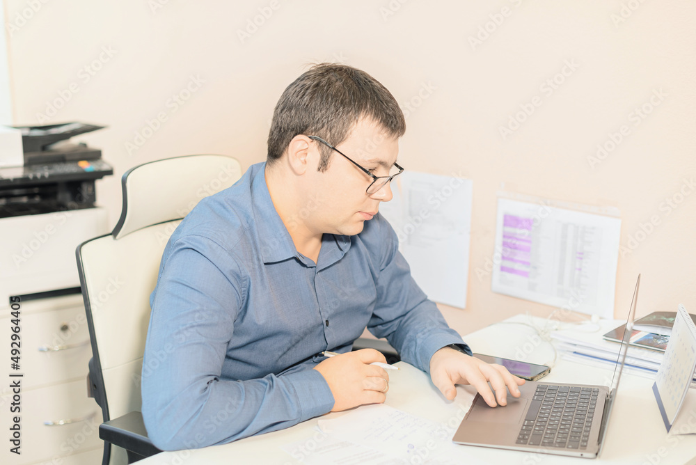 Man employee in blue shirt and glasses sitting at the desk in office using laptop computer. Business, busy working day concept Man employee in blue shirt and glasses sitting at the desk in office using laptop computer. Business, busy working day concept