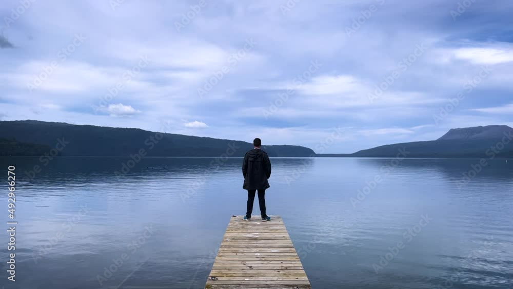 A young caucasian man stands on a boat jetty on the banks of Lake Tarawera in New Zealand looking off towards the horizon A young caucasian man stands on a boat jetty on the banks of Lake Tarawera in New Zealand looking off towards the horizon