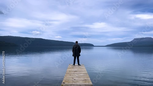 A young caucasian man stands on a boat jetty on the banks of Lake Tarawera in New Zealand looking off towards the horizon photo