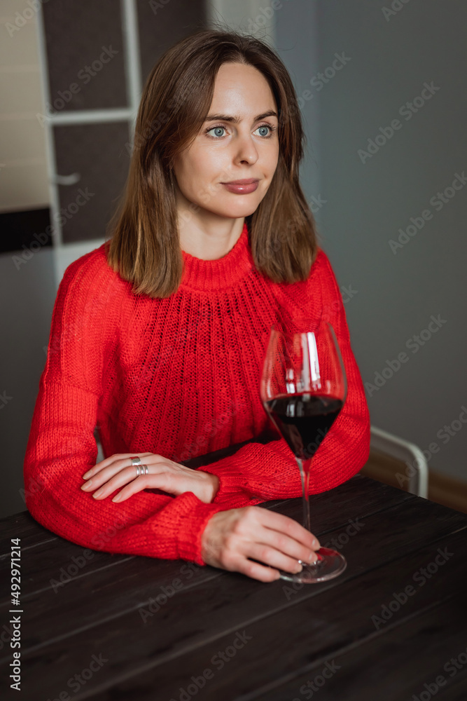 A pretty young caucasian woman sitting at the wooden table in the kitchen with a glass of red wine in her hands A pretty young caucasian woman sitting at the wooden table in the kitchen with a glass of red wine in her hands