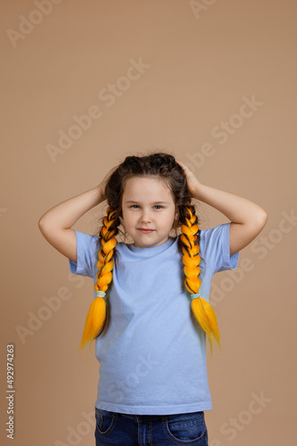 Offended, little misbehaving girl holding head with hands looking camera with sullen look having kanekalon braids of yellow color in blue t-shirt on beige background.