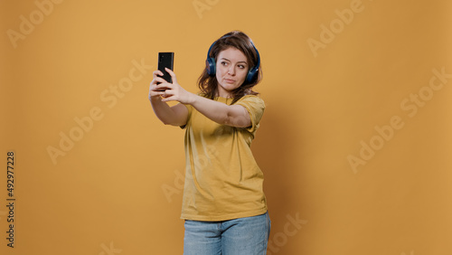 Casual woman taking a selfie using mobile phone while listening to music on wireless headphones in studio. Young person using smartphone to take picture using front camera while wearing earphones.