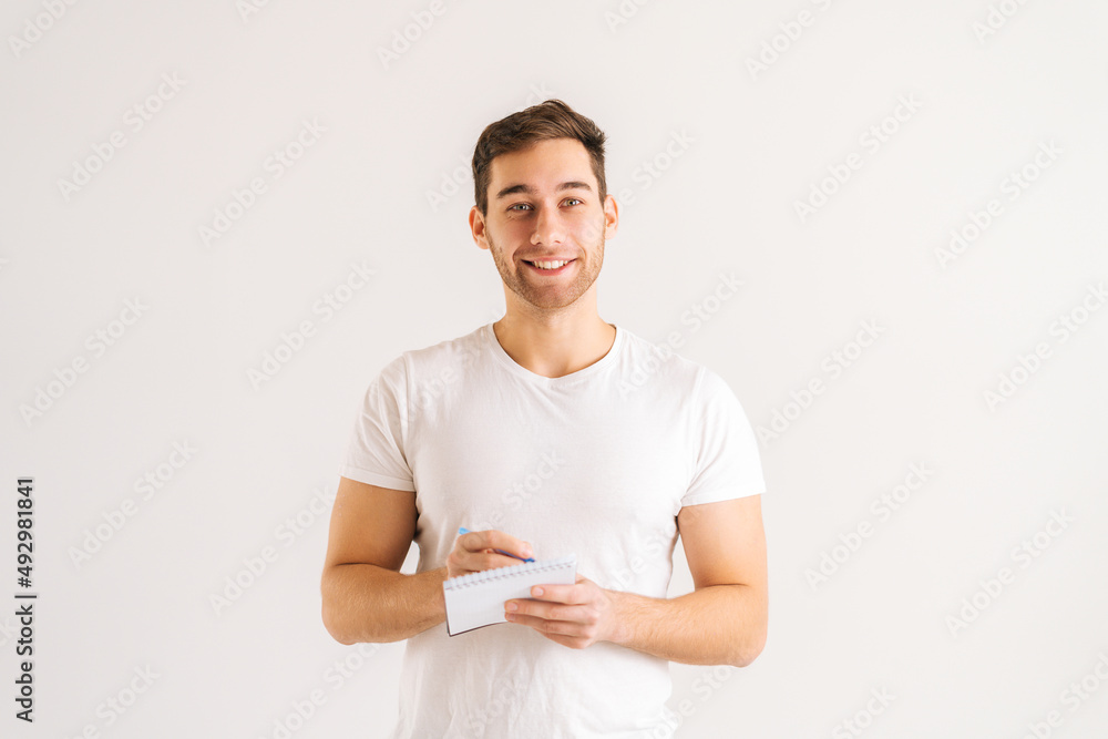 Studio portrait of cheerful young man writing in copybook with pen standing on white isolated background. Happy male student studying, preparing for exam, making notes in paper notebook. Studio portrait of cheerful young man writing in copybook with pen standing on white isolated background. Happy male student studying, preparing for exam, making notes in paper notebook.