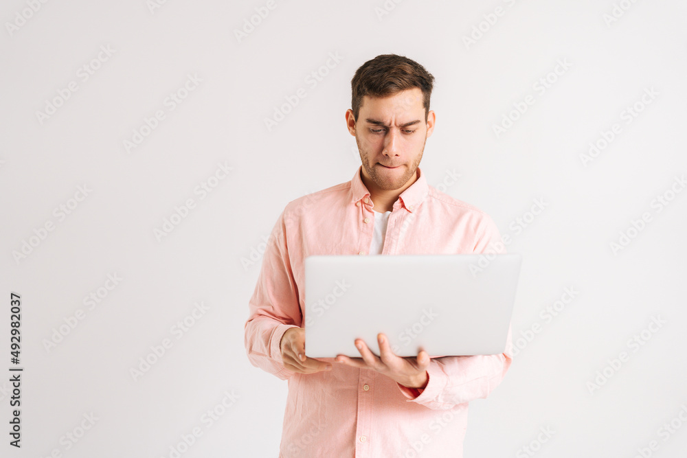 Portrait of serious young man standing with laptop and typing on keyboard, doing freelance job, writing email on white isolated background. Studio shot of pensive male posing with pc looking at screen Portrait of serious young man standing with laptop and typing on keyboard, doing freelance job, writing email on white isolated background. Studio shot of pensive male posing with pc looking at screen