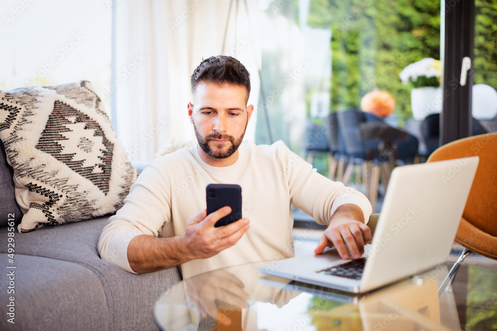 Serious faced man text messaging and using laptop while working from home Serious faced man text messaging and using laptop while working from home