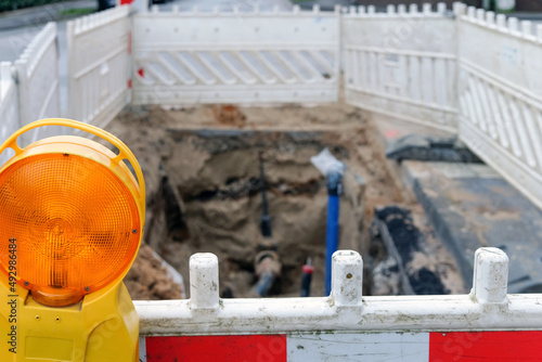 Offene Baugrube mit Wasseranschluss und anderen Versorgungsleitungen im unscharfen Hintergrund in einer Straße, mit Absperrbarken, Schutzgitter und Baustellenlampe im Vordergrund, selektiver Fokus