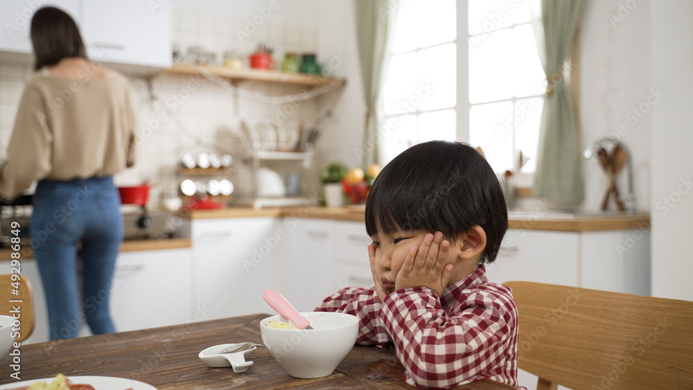 selective focus of upset Asian preschool boy holding face with a look of hate as he picks up vegetable from his bowl at dining table. his mother is cooking in kitchen at background selective focus of upset Asian preschool boy holding face with a look of hate as he picks up vegetable from his bowl at dining table. his mother is cooking in kitchen at background