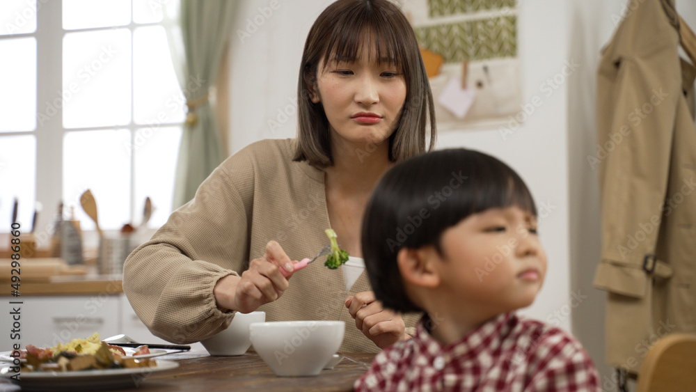 frustrated Asian mother holding a fork with vegetable and asking his baby son to take a bite in dining room at home. the boy turns head away and refuses to eat frustrated Asian mother holding a fork with vegetable and asking his baby son to take a bite in dining room at home. the boy turns head away and refuses to eat