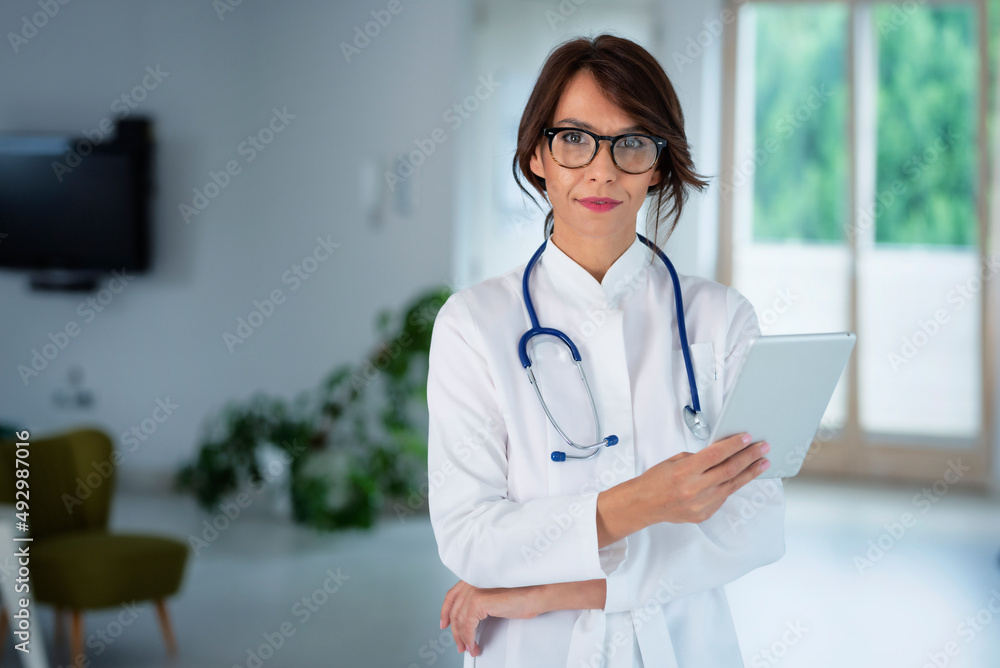 Attractive female doctor portrait while holding tablet in her hand and standing at the hospital corridor