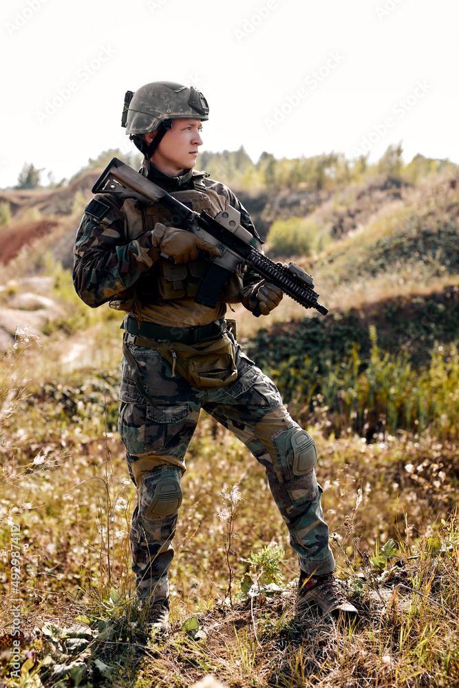 Portrait Of Serious Caucasian Man In Military Combat Uniform, Looking At Side Holding Weapon Rifle Gun In Hands, Standing Alone In Field, Ready To Shoot. Military Forces concept