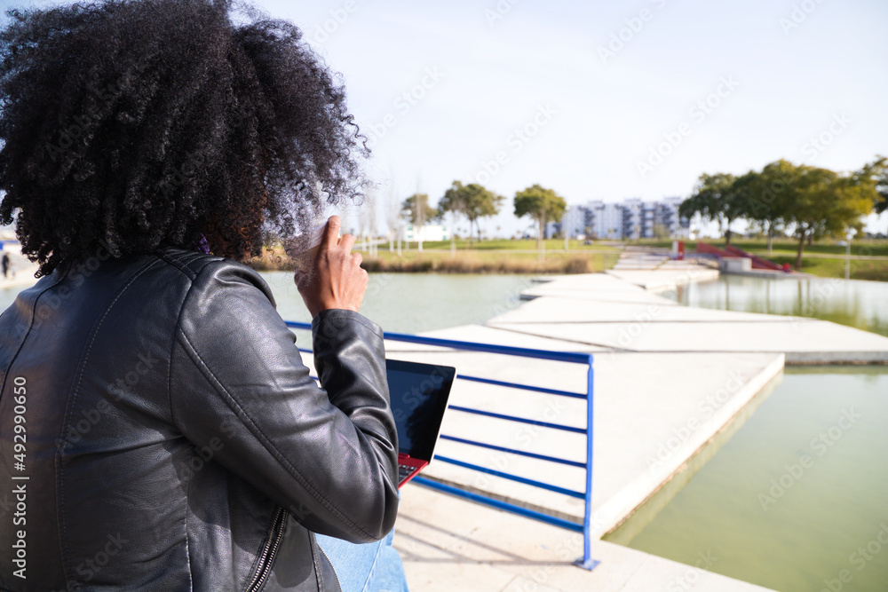 Young and beautiful Afro-American woman with laptop computer shopping online, checking her mail, social networks and working in the park. The woman is wearing casual clothes and drinking coffee, tea.