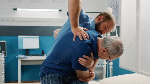 Osteopath helping senior man to crack back bones in cabinet, doing physical therapy exercise. Male assistant using physiotherapy procedure to increase mobility and treat patient.
