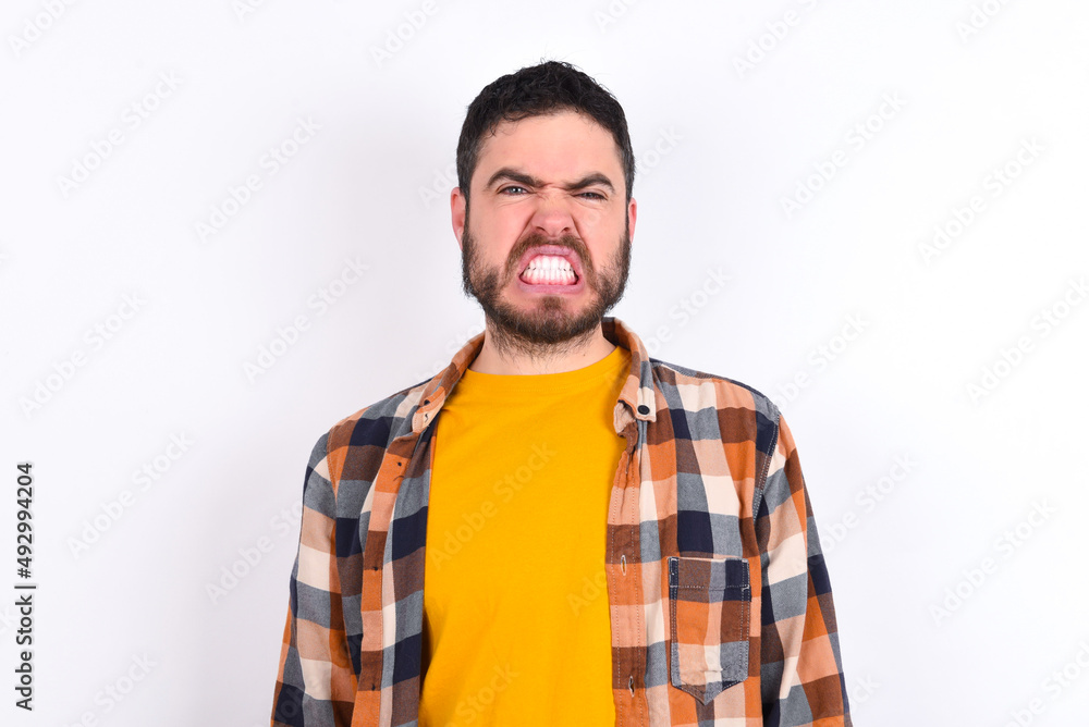 Mad crazy young caucasian man wearing plaid shirt over white background clenches teeth angrily, being annoyed with coming noise. Negative feeling concept. Mad crazy young caucasian man wearing plaid shirt over white background clenches teeth angrily, being annoyed with coming noise. Negative feeling concept.