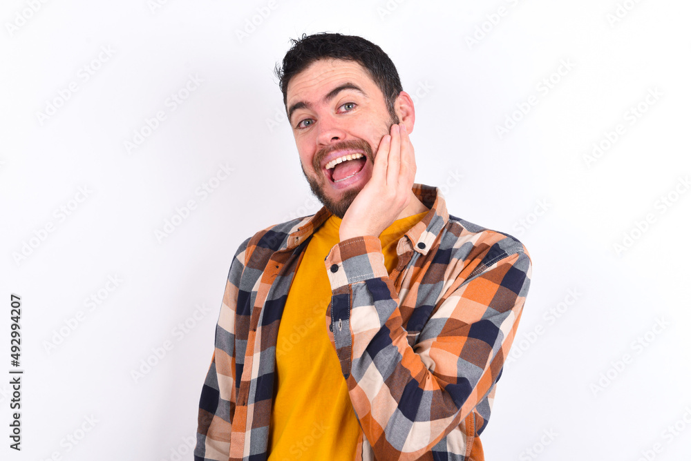 Shocked, astonished young caucasian man wearing plaid shirt over white wall looking surprised in full disbelief wide open mouth with hand near face. Positive emotion facial expression body language. Shocked, astonished young caucasian man wearing plaid shirt over white wall looking surprised in full disbelief wide open mouth with hand near face. Positive emotion facial expression body language.