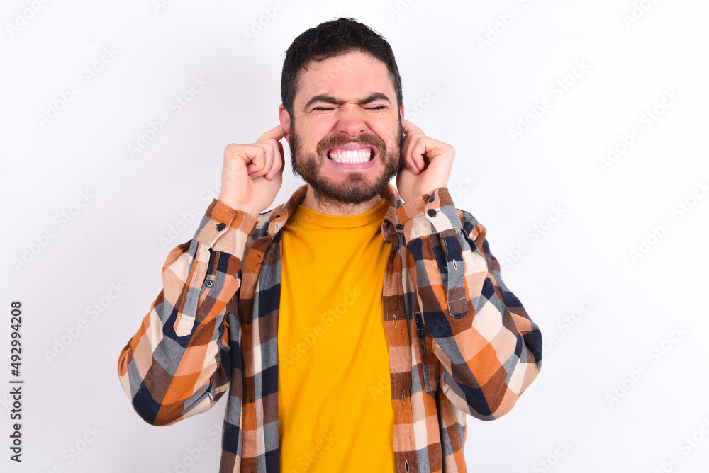 Stop making this annoying sound! Unhappy stressed out young caucasian man wearing plaid shirt over white background making worry face, plugging ears with fingers, irritated with loud noise. Stop making this annoying sound! Unhappy stressed out young caucasian man wearing plaid shirt over white background making worry face, plugging ears with fingers, irritated with loud noise.