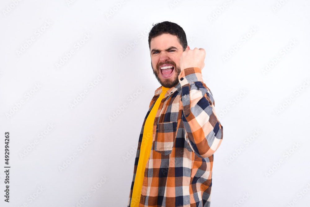 Fototapeta premium Overjoyed young caucasian man wearing plaid shirt over white background glad to receive good news, clenching fist and making winning gesture.