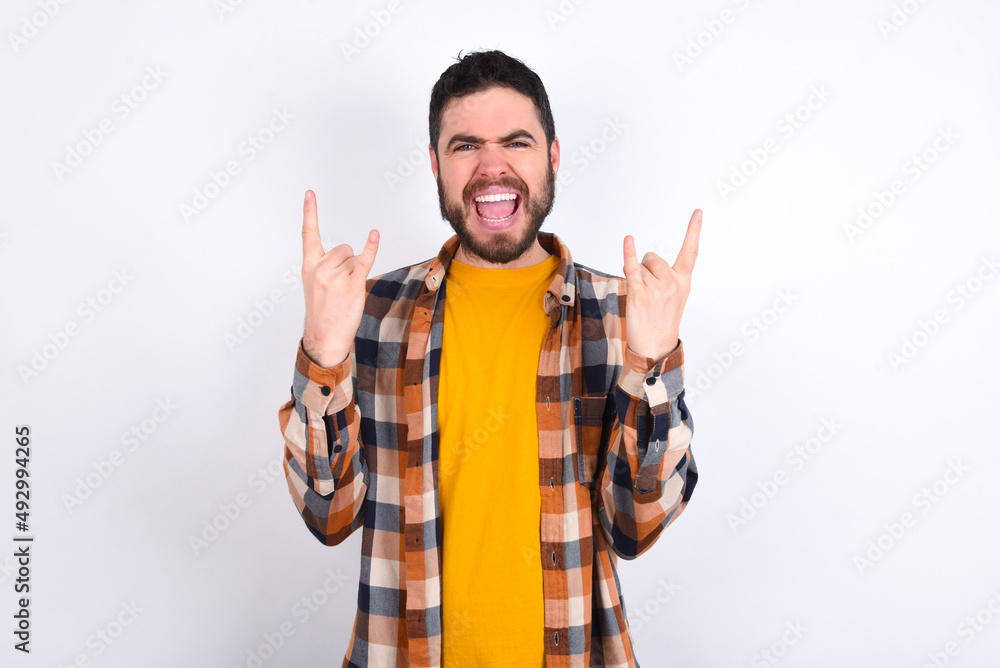 Born to rock this world. Joyful young caucasian man wearing plaid shirt over white background screaming out loud and showing with raised arms horns or rock gesture. Born to rock this world. Joyful young caucasian man wearing plaid shirt over white background screaming out loud and showing with raised arms horns or rock gesture.