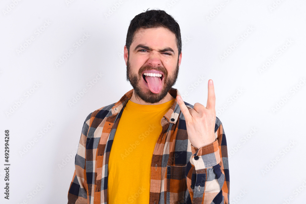 Fototapeta premium Portrait of a crazy young caucasian man wearing plaid shirt over white background showing tongue horns up gesture, expressing excitement of being on concert of band.