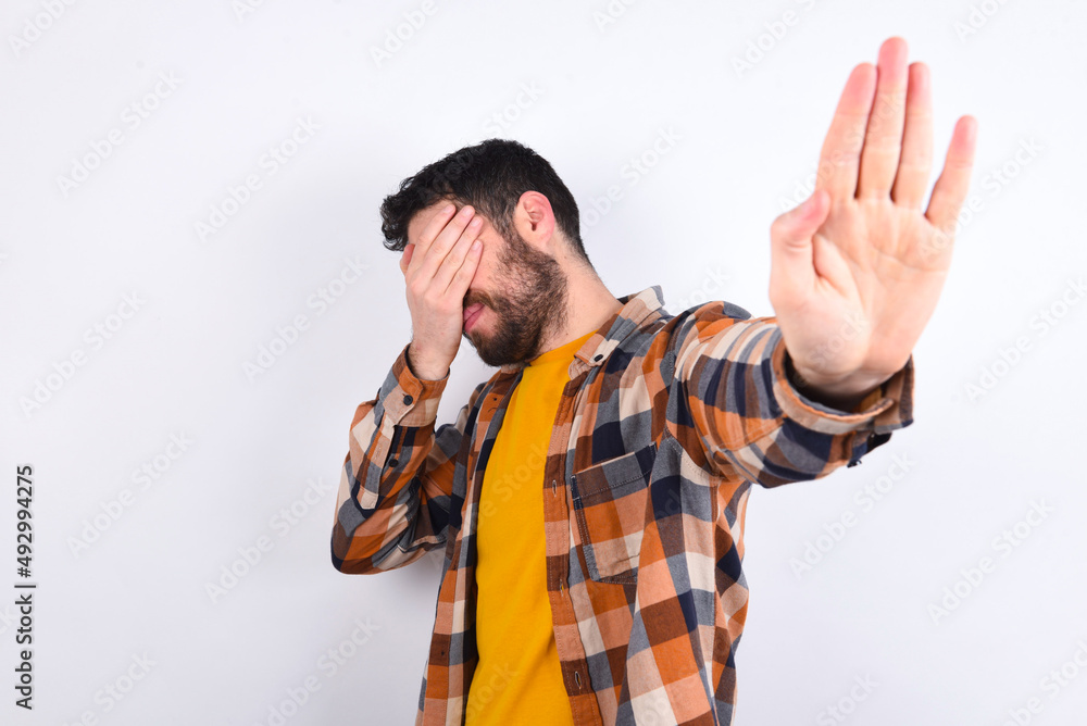 Naklejka premium young caucasian man wearing plaid shirt over white background covers eyes with palm and doing stop gesture, tries to hide. Don't look at me, I don't want to see, feels ashamed or scared.