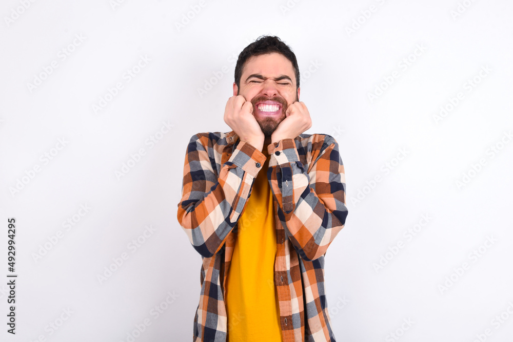 Portrait of young caucasian man wearing plaid shirt over white background being overwhelmed, expressing excitement and happiness with closed eyes and hands near face. Portrait of young caucasian man wearing plaid shirt over white background being overwhelmed, expressing excitement and happiness with closed eyes and hands near face.