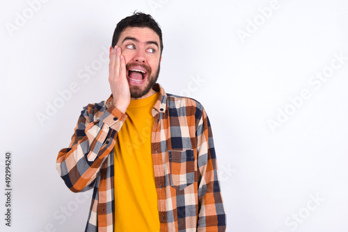 young caucasian man wearing plaid shirt over white background excited looking to the side hand on face. Advertisement and amazement concept.