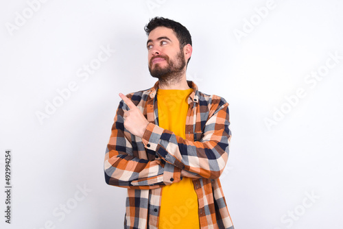 Portrait of young caucasian man wearing plaid shirt over white background posing on camera with tricky look, presenting product with index finger. Advertisement concept.