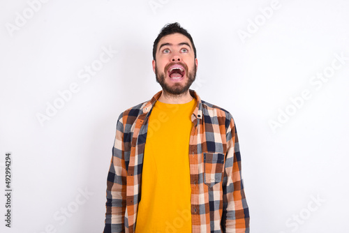 Surprised young caucasian man wearing plaid shirt over white background, shrugs shoulders, looking sideways, being happy and excited. Sudden reactions concept.