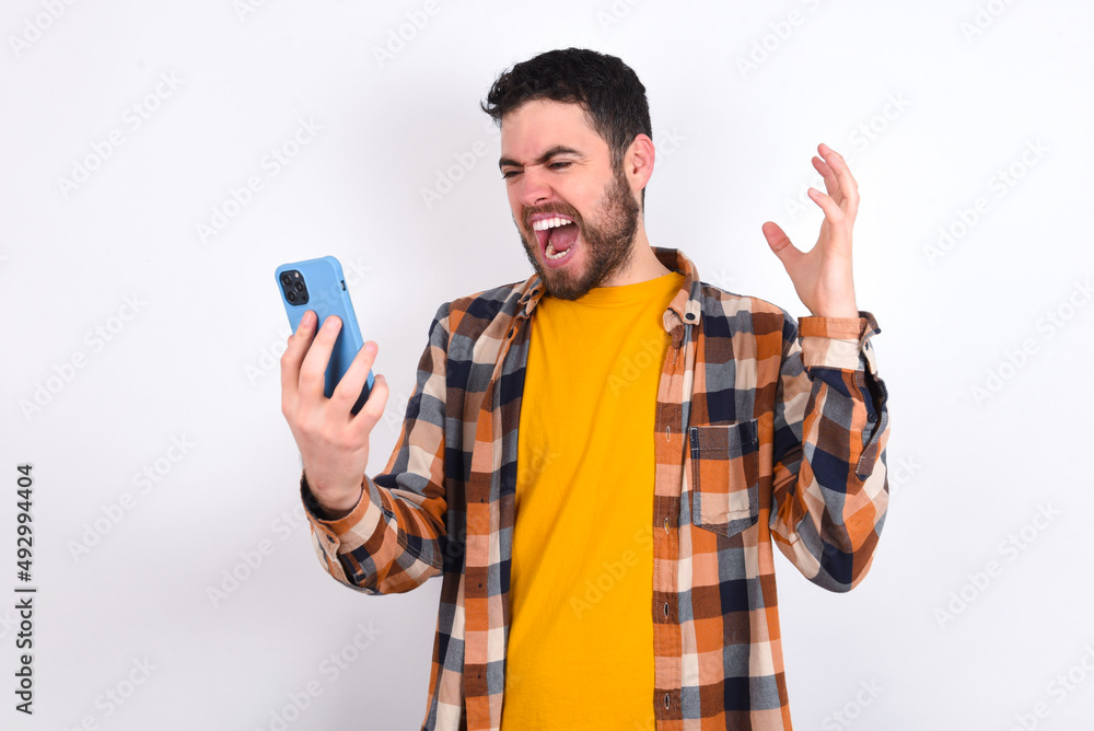 Photo of outraged annoyed young caucasian man wearing plaid shirt over white background holds cell phone, makes call, argues with colleague, expresses negative emotions. People and anger. Photo of outraged annoyed young caucasian man wearing plaid shirt over white background holds cell phone, makes call, argues with colleague, expresses negative emotions. People and anger.