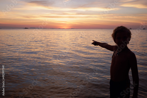 Silhouette of the boy shows his hand to the horizon and sunset at the sea. Lifestyle, Childhood, Travel, Freedom concept. an interested child theme. Beautiful landscape